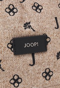 Tissu beige tissé avec des motifs floraux abstraits noirs et de lettres, et un patch en cuir noir portant l'inscription « JOOP ! » en lettres argentées.
