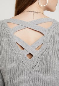 Grå stickad tröja med korsad ryggdesign och slitsar. Texturen är lös och öppen, med en V-halsringning och inga ytterligare dekorativa inslag.