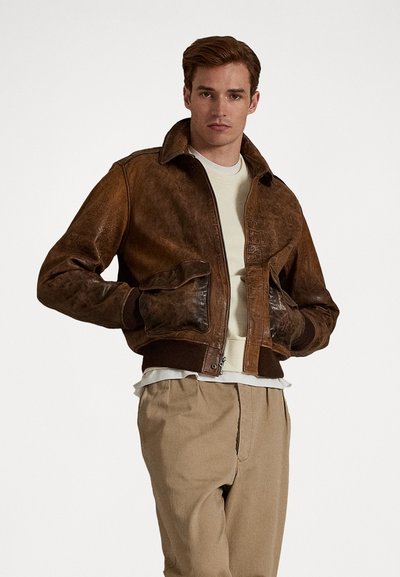 Jaqueta bomber de couro castanho com gola, punhos canelados e fecho de correr. Apresenta mangas texturizadas e bolsos frontais, usada sobre uma camisa clara.