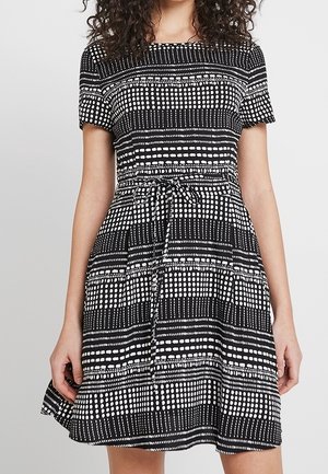 Femme portant une robe noire à manches courtes avec des motifs géométriques à pois blancs et rayures, cintrée à la taille par une ceinture nouée.