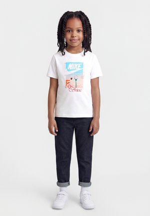 Ragazza con capelli intrecciati, indossa una t-shirt bianca Nike con grafica da tennis e testo "Rise and Shine", jeans scuri, calze bianche e scarpe da ginnastica.