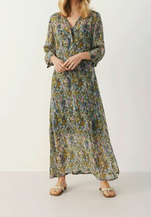 Femme portant une longue robe à imprimé floral avec des manches trois-quarts et des sandales plates tressées beige, debout devant un fond uni.