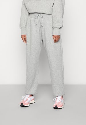 Personne portant un pantalon de survêtement gris clair avec taille à cordon et des baskets rose et blanc, debout devant un fond clair uni.
