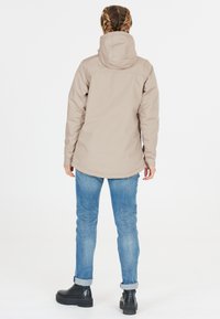 Beige vattentät jacka med huva, rak skärning och slät textur, bärs över blå jeans och svarta ankelskor.