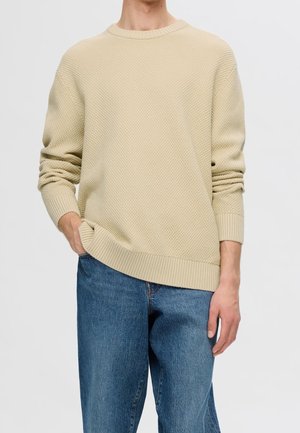 Personne portant un pull en maille beige clair texturé et un jean en denim bleu, debout avec une main dans la poche et l'autre détendue.