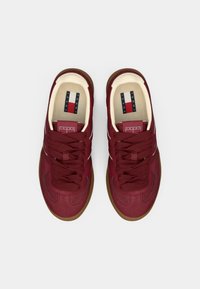 Červené tenisky s textúrovaným povrchom, tmavými šnúrkami, bielym pruhom na ozdobu a gumovou podrážkou. Logo umiestnené na jazyku.