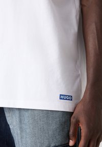 Balta kokvilnas T-kreklu ar zilu "HUGO" logo apakšmalā; īsas piedurknes, brīvs piegriezums, gluda tekstūra, valkāta ar denima biksēm.