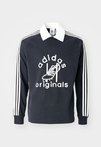 Polo shirt negru cu mânecă lungă, cu guler alb și trei dungi albe pe mâneci, având o grafică mare albă a unei ghete de fotbal și textul "adidas originals."