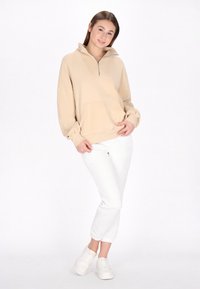 Beige dragkedja sweatshirt med ståkrage, framficka och ribbade ärmslut, i kombination med vita smalrandiga byxor och vita sneakers.