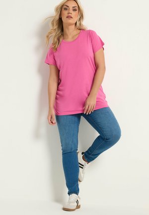 Kvinde med langt blondt hår iført en lyserød kortærmet skjorte, blå jeans og hvide sneakers, læner sig op ad en hvid væg.