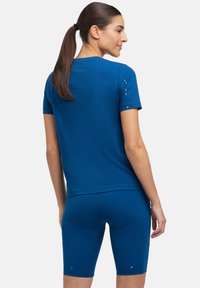 Maglietta sportiva blu con maniche corte, dalla texture liscia, abbinata a pantaloni corti blu coordinati. Accenti notevoli includono piccole lettere stampate sulle maniche.