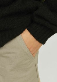 Fekete kötött pulóver ribbelt mandzsettákkal világos bézs nadrággal; a kéz egy oldazsebben pihen, bemutatva a szövet textúráját és varrásait.