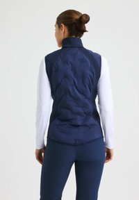 Marinblå vadderad puffer-väst med hög krage, strukturerad yta och elastiska sidor. Bärs över en långärmad vit topp och marinblå byxor.