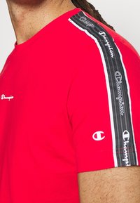Červená košile s krátkým rukávem s černobílým pruhem s logem Champion na rameni a rukávu, nošená osobou s viditelnou pletí a vlasy.