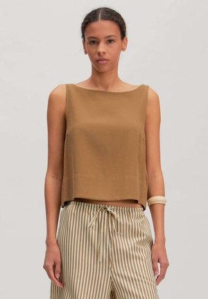 Vrouw draagt een mouwloze bruine top, gestreepte beige en witte broek met aantrekkoord en een beige armband aan haar linkerarm, staand tegen een effen achtergrond.