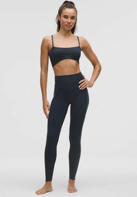 Nő mezítláb áll, fekete magas derekú leggingsben és fekete crop topban, az egyik kezét a csípőjére téve pózol szürke háttér előtt.