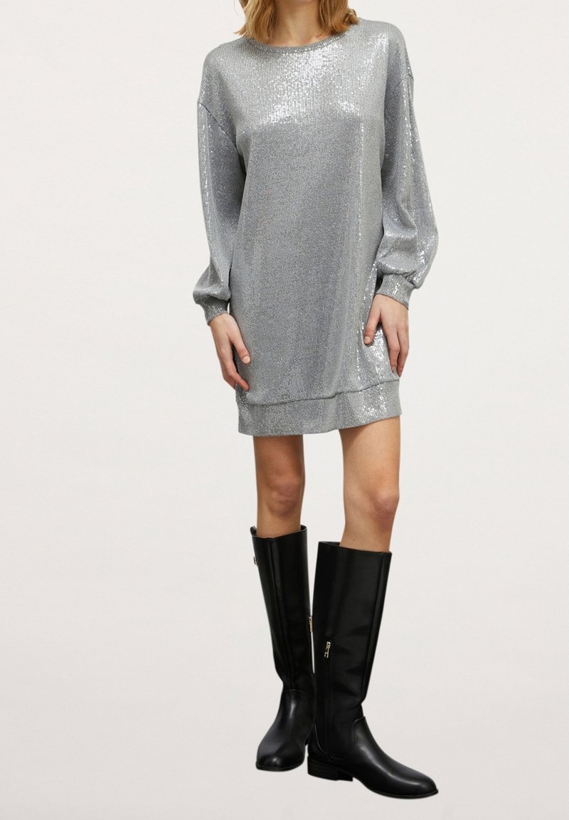 Femme portant une mini-robe à manches longues en sequins argentés associée à des bottes en cuir noires montant jusqu'aux genoux, sur un fond uni.