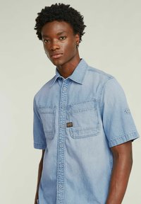 Ljust blå kortärmad jeansskjorta med knappslå, två framfickor och en logotypetikett på bröstet. Mjuk textur, avslappnad design.