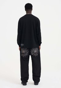 Zwarte longsleeve shirt met losse zwarte broek, voorzien van spinnenwebborduursel op de achterzakken en subtiele logo-details.