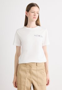 T-shirt bianca in cotone con maniche corte; presenta un logo stampato in grigio scuro. Abbinata a pantaloni beige, il modello è in piedi su uno sfondo neutro.