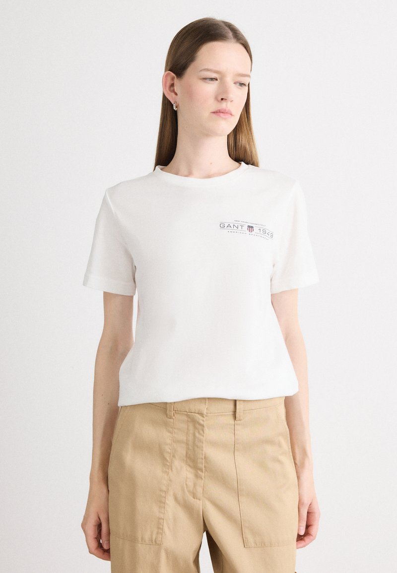 T-shirt bianca in cotone con maniche corte; presenta un logo stampato in grigio scuro. Abbinata a pantaloni beige, il modello è in piedi su uno sfondo neutro.