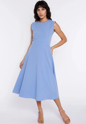 Kobieta o ciemnych włosach, ubrana w bezrękawnikową, jasnoniebieską sukienkę do połowy łydki w stylu A-linii oraz złote sandały na wysokim obcasie, stojąca z jedną ręką na biodrze.