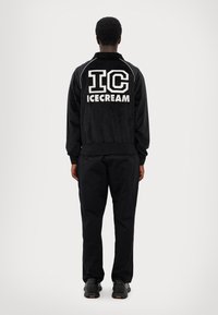 Veste en velours noir avec un grand texte blanc "IC ICECREAM" dans le dos, des poignets et un col côtelés ; associée à un pantalon noir et des baskets noires.