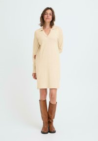 Abito a maglia beige con scollatura a V profonda e maniche lunghe, caratterizzato da una vestibilità comoda e una finitura leggermente texturizzata, abbinato a stivali marroni al ginocchio.