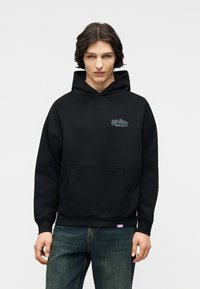 Mlad moški s srednje dolgimi temnimi lasmi, oblečen v črno kapuco z majhnim belim napisom na prsih in modre kavbojke, stoji pred belim ozadjem.