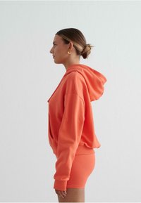 Koraaloranje cropped hoodie met een trekkoord, voorzien van een buidelzak, gecombineerd met bijpassende strakke shorts. Stevige textuur op de stof.