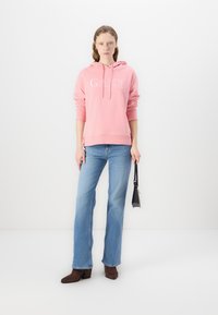 Rosa hoodie med dragsko och logotyp, matchad med ljusblå utsvängda jeans och bruna högklackade stövlar. Håller en liten svart väska.