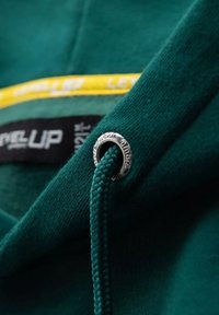 Tessuto del cappuccio verde con un accento giallo e dettagli con occhielli argentati. Presenta un cordoncino intrecciato e un'etichetta con il marchio "LEVEL UP".