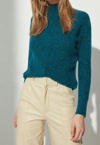 Tyrkysový pletený svetr s vysokým výstřihem, texturovaným vzorem a volným střihem, kombinovaný s krémovými kalhotami z umělé kůže.