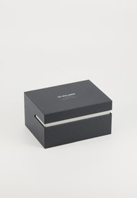Boîte rectangulaire noire avec une surface texturée, présentant une bande blanche et "DI MILANO" imprimé sur le dessus en argent. Design fermé.
