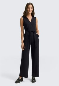 Une combinaison noire avec un design enveloppant, un décolleté en V profond, des jambes larges, une taille à nouer et des poches latérales, associée à des chaussures noires à enfiler.