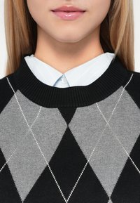 Fekete és szürke argyle mintás pulóver, bordás kerek nyakkal, egy világoskék gombos ingre rétegezve, amelyen vékony függőleges csíkok találhatók.