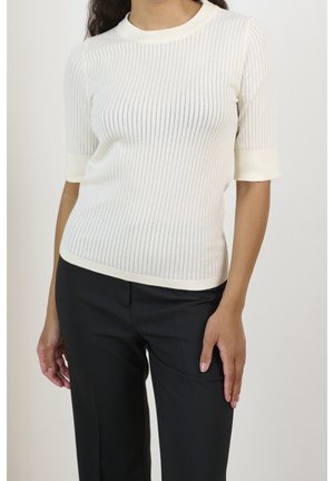 Femme portant un pull blanc à manches courtes côtelé et un pantalon noir sur mesure, debout avec une main reposant sur sa cuisse.