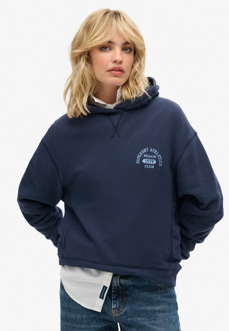 Felpa con cappuccio blu navy realizzata in materiale morbido, con un logo frontale in azzurro. Vestibilità ampia con orlo e polsini a costine, indossata sopra una maglietta bianca.