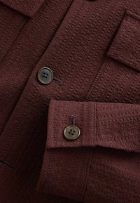 Gros plan sur un tissu marron foncé texturé avec deux boutons marron sur une manchette et un rabat de poche.