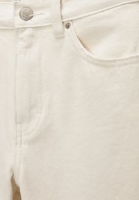 Gros plan sur la ceinture d’un jean en denim couleur crème avec un bouton métallique et les détails des coutures de la poche avant.