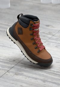 Botte de randonnée marron et brun foncé avec des lacets rouges, col rembourré noir autour de la cheville et semelle texturée sur un sol en pierre gris clair.