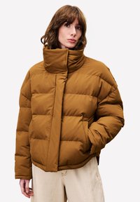 Vrouw met bruin golvend haar, gekleed in een hoge kraag, bruine pufferjack en lichtbeige broek, staan met één hand in de zak.