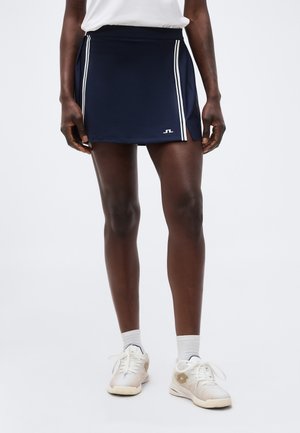 Γυναίκα που φορά μπλε αθλητική φούστα με λευκές ρίγες στα πλαϊνά, λευκές κάλτσες και λευκά αθλητικά παπούτσια, στέκεται μπροστά σε ένα απλό φόντο.