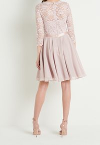 Woman wearing pale pink lace top dress with three-quarter sleeves, pleated skirt, and matching high-heeled ankle-strap shoes, standing back view.