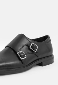 Chaussures en cuir noir avec deux boucles en argent, finition lisse, bout arrondi et un faible talon solide. Design simple et épuré.