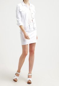 Giacca di jeans bianca con un design aderente e bottoni a pressione argentati, abbinata a una minigonna bianca. Sandali con zeppa con cinturini bianchi e tacco in legno.