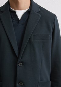 Marineblauwe textuur blazer met een notch lapel, twee knopen en een opgestikte zak. Geddragen over een donker V-halstrui.