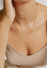 Collares en tono dorado de capas que presentan cadenas delicadas con pequeños acentos de cuentas. Llevados sobre una blusa de color neutro y sólido. Textura suave.