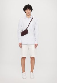 Lys blåstripet skjorte med knapp, hvite shorts og en liten burgunder crossover-veske. Modellen har på seg hvite sneakers og står mot en ensfarget bakgrunn.