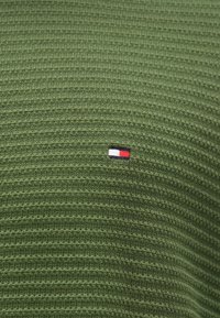 Ελαιόχρωμο πλεχτό πουλόβερ με ριγωτή υφή και μικρό κεντημένο λογότυπο σε κόκκινο, λευκό και μπλε στη αριστερή πλευρά.